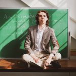 Aandeg Muldrew sits cross-legged on top of a desk in front of a green chalkboard.