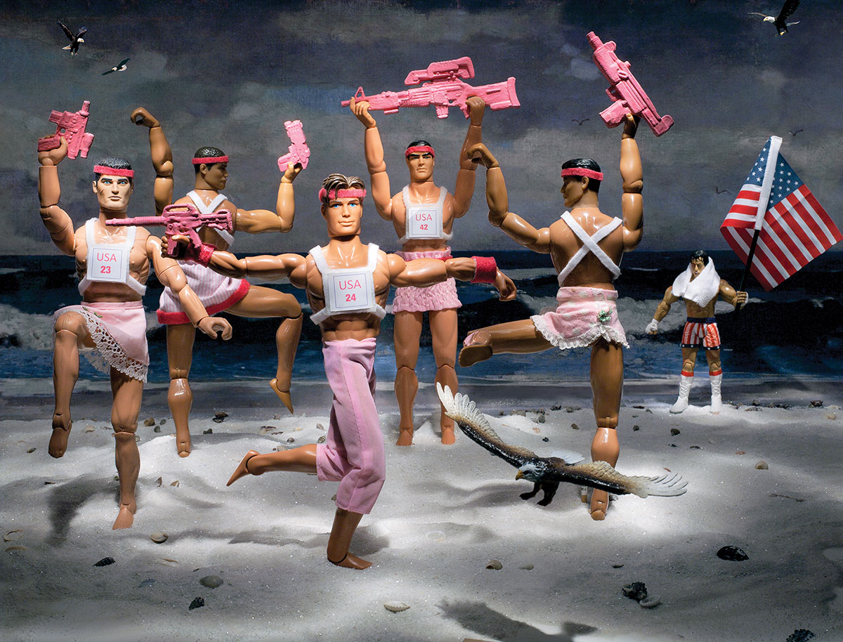 A team of toy action figures with pink clothing and guns dance on a beach, with eagles flying around and a man with an American flag in the background.