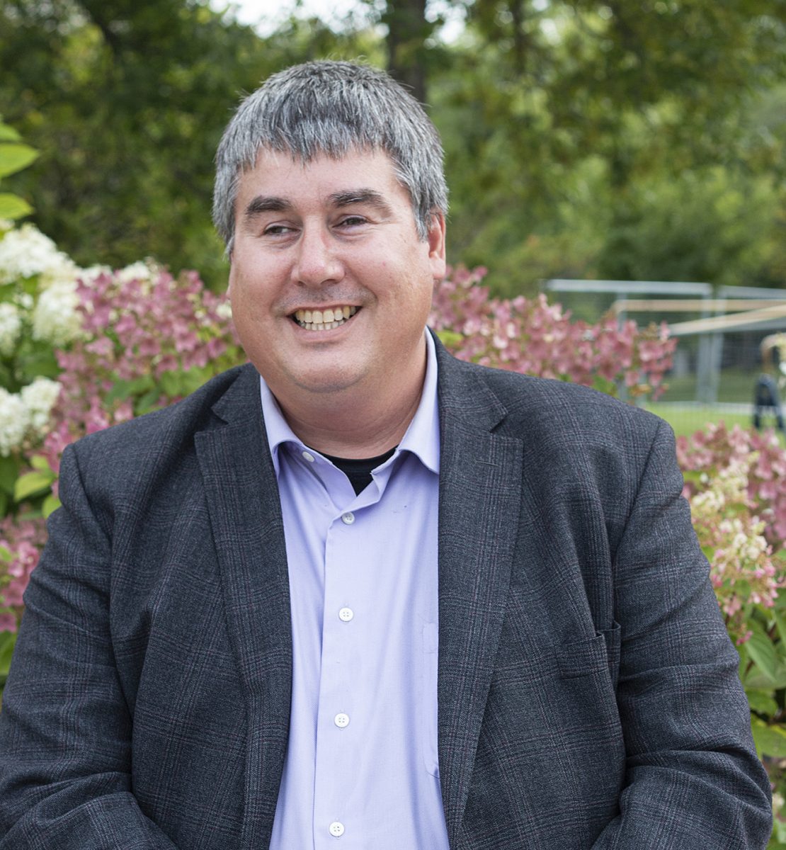 Todd Duhamel sitting in front of flowers and shrubs on the campus of the University of Manitoba