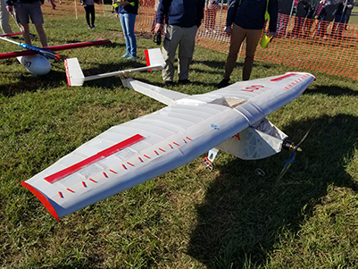 A white model aircraft with about 8 foot wing span sits on green grass. The wings and tail section have red accents. 