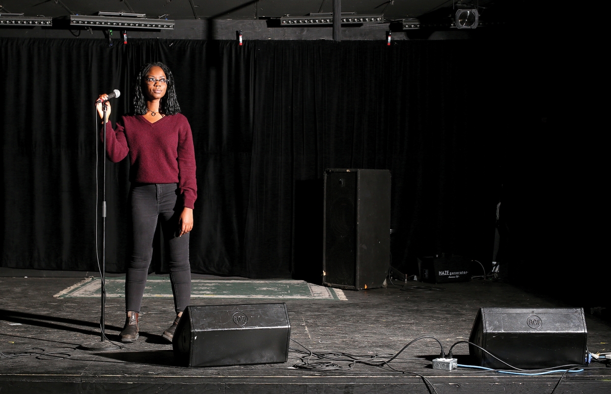 Chimwemwe Undi stands next to a microphone on a dimly lit stage.