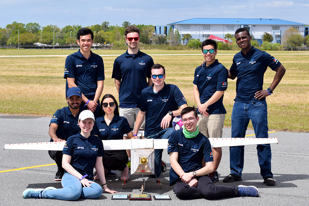 Nine students wearing team branded navy blue polo shirts stand/sit posing facing the camera. They are surrounding their aircraft build which is white and about 8 feet in wing span. In front of the plane are three award plaques.
