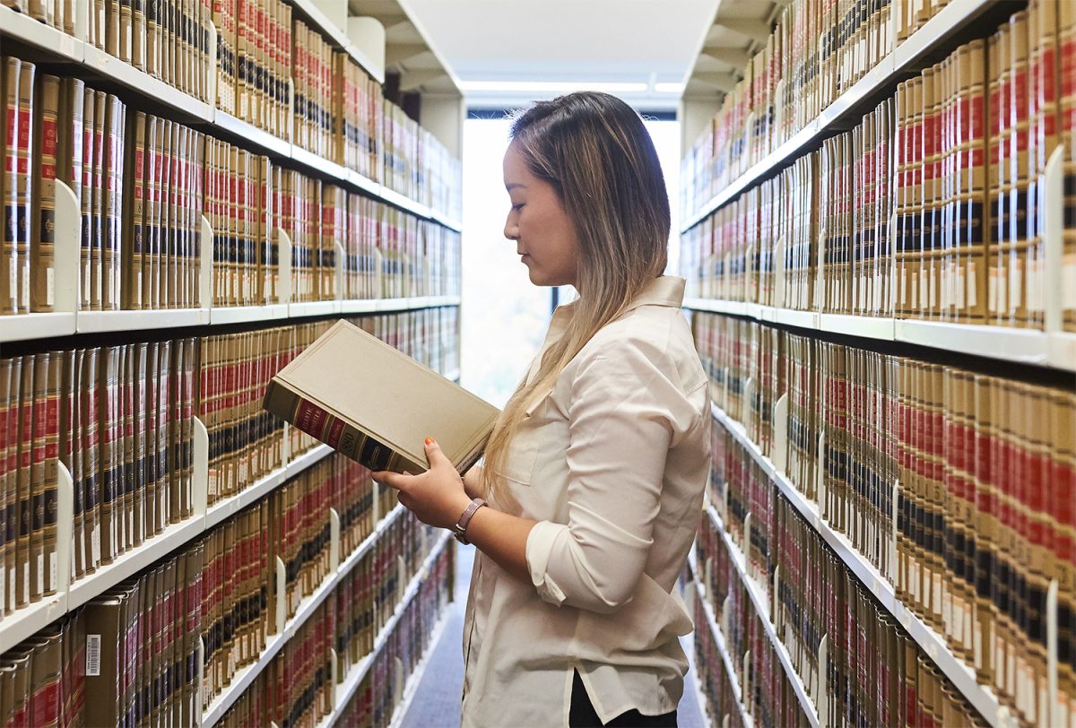 A law student holds a book in her hand standing in a long row of library shelves full of books.