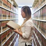 A law student holds a book in her hand standing in a long row of library shelves full of books.