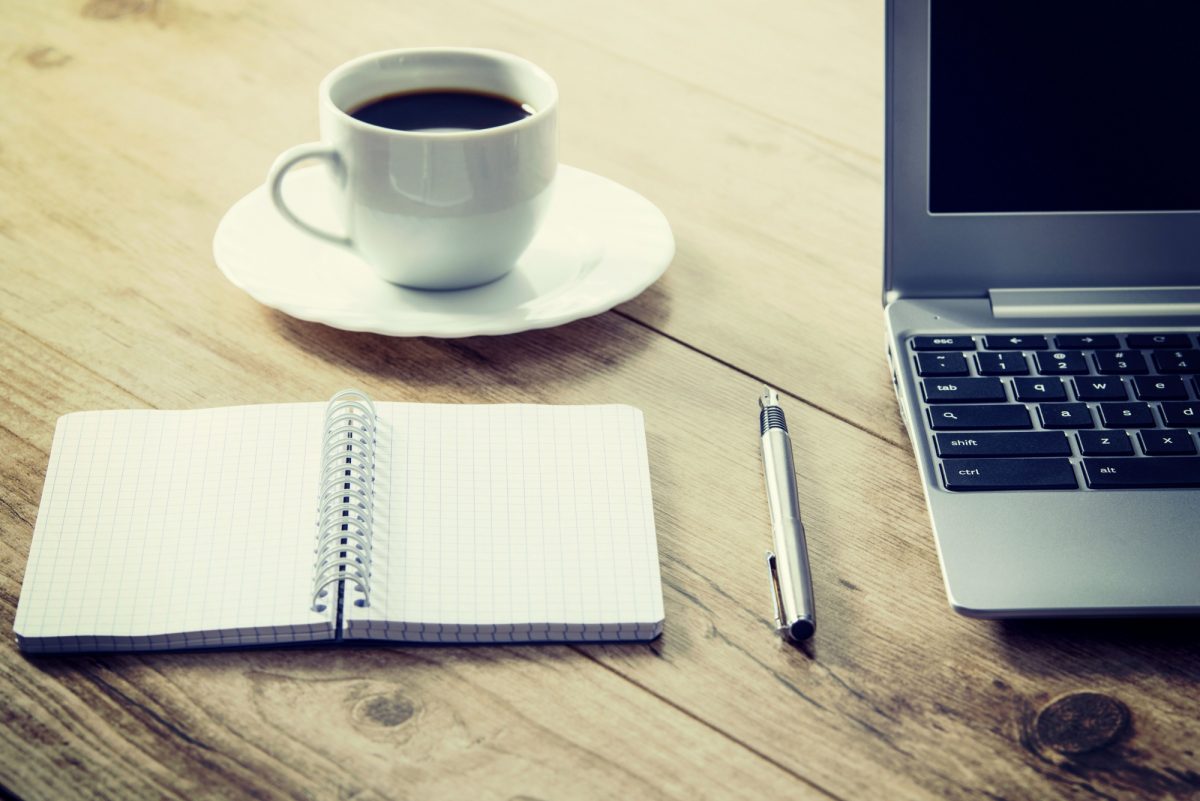 Cup of coffee on a saucer sits on a wooden tabletop beside a laptop, pen, and open notebook.