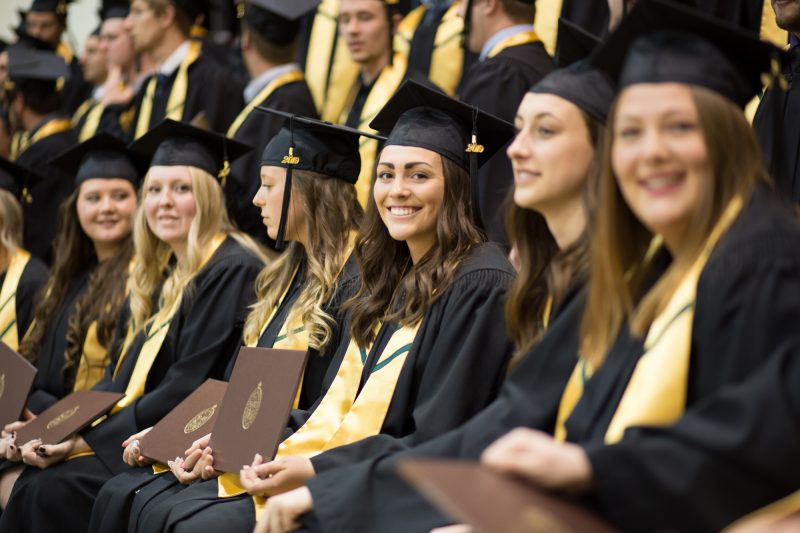 A group of UM grads sit in a row at Convocation. They wear black caps and gowns with golden hood linings. The graduate in the middle smiles at the camera as all hold up their degree parchments.
