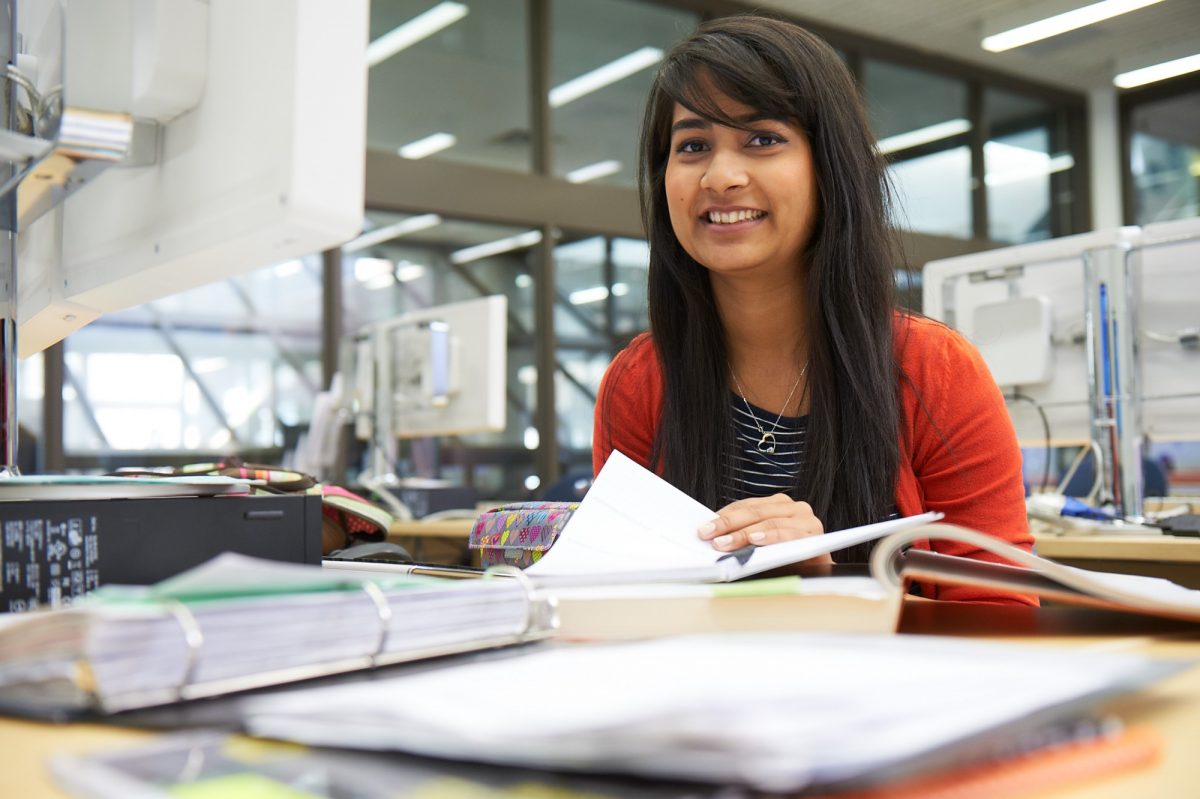 Woman with dark hair smiling in the Asper School of Business Drake Building. She has books and papers infront of her that are out of focus.