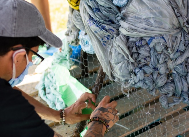 A man wearing a cap, glasses, and a mask works on an art project made of out of different plastics like plastic bags.
