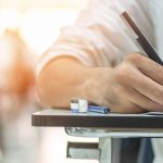 A person sits at a desk, writing on a paper with pen.