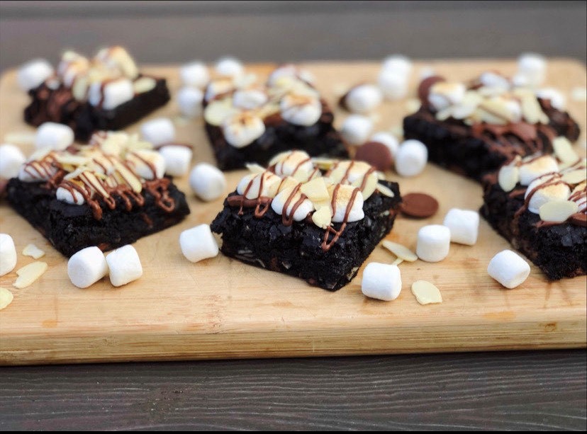 Six dark brownies with marshmallows and drizzle on top. They are sitting on a wooden platter. Brownies made by Babes Brownies.