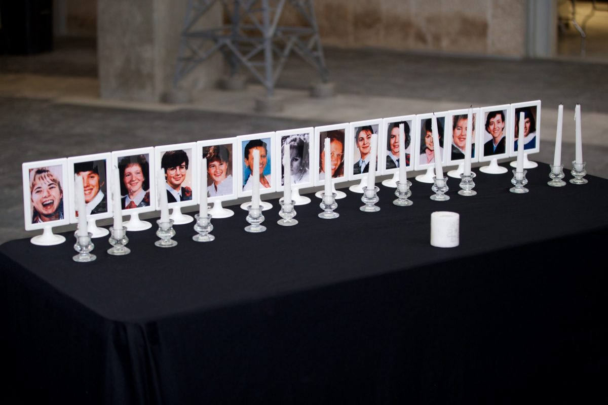 A table covered with a black table cloth. On top on the table fourteen photo frames with photos of each of the victims, all of whom are young women. In front of each of the photos is a candle.