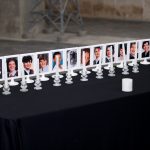 A table covered with a black table cloth. On top on the table fourteen photo frames with photos of each of the victims, all of whom are young women. In front of each of the photos is a candle.