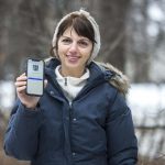 Doctor Zahra Moussavi standing outside smiling and posing for the camera while holding up a smartphone.