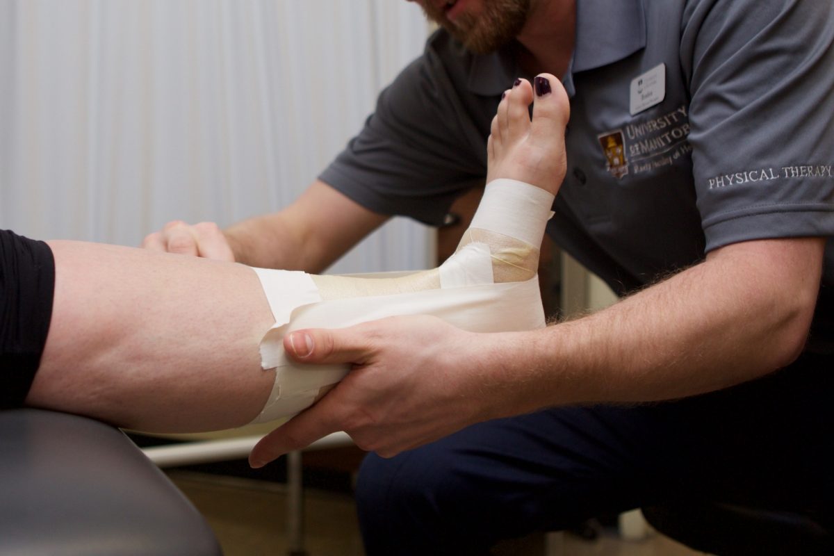 The arms and upper body of a physiotherapy student are seen. The student is taping a woman's bare foot and ankle with athletic tape.