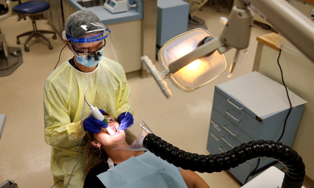 A dental student wearing personal protective equipment performs a dental procedure on a patient while a plastic funnel attached to plastic tubing vacuums aerosols away from the patient's chin area.