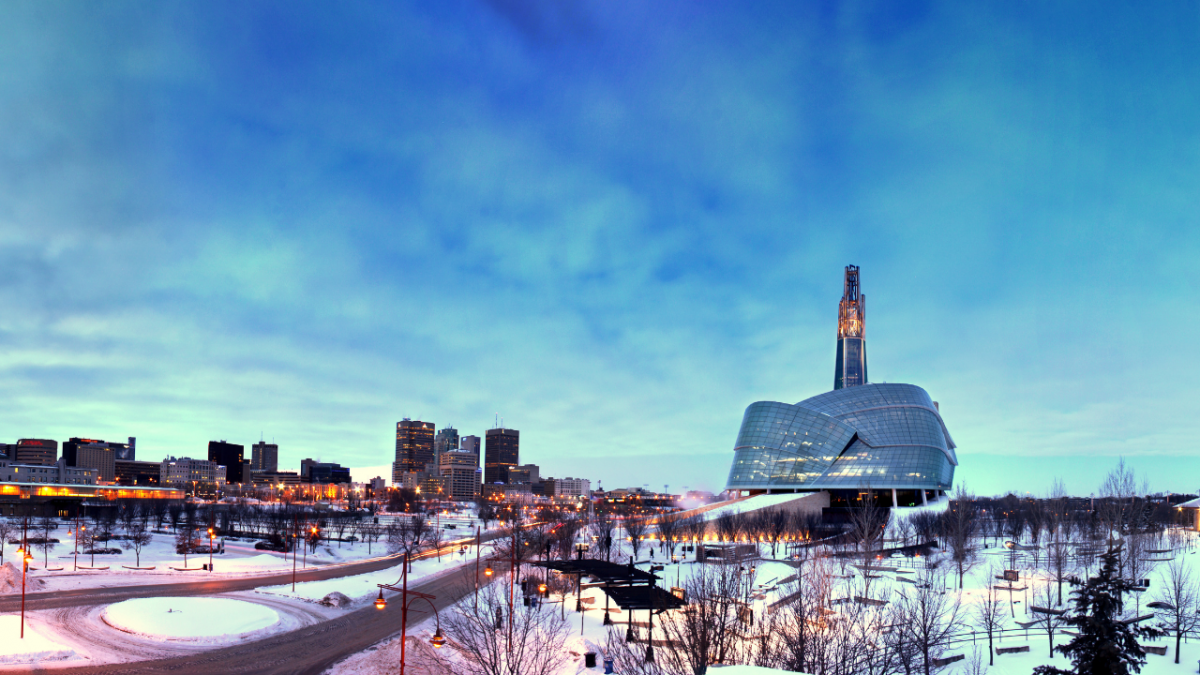 Canadian Museum of Human Rights building with a blue sky background, with a snowy foreground and the downtown skyline in the distance.