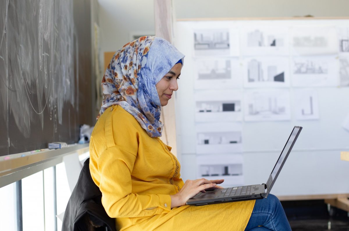 A woman in a yellow shirt and a blue and orange head scarf works on a laptop in her lap.