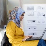 A woman in a yellow shirt and a blue and orange head scarf works on a laptop in her lap.