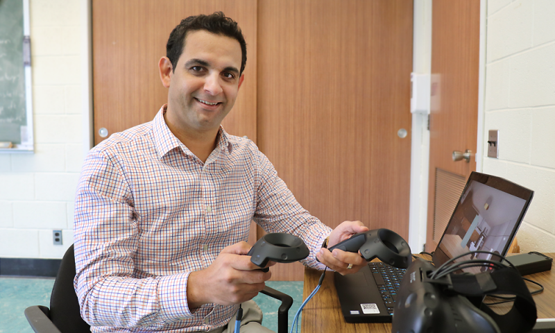 Dr. Amine Choukou holds a controller in each hand while sitting at a desk. A laptop computer is open on the desk.