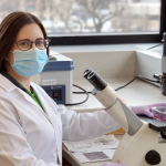 Dr. Deanna Santer, wearing a white lab coat and a surgical mask, sits in front of a microscope.