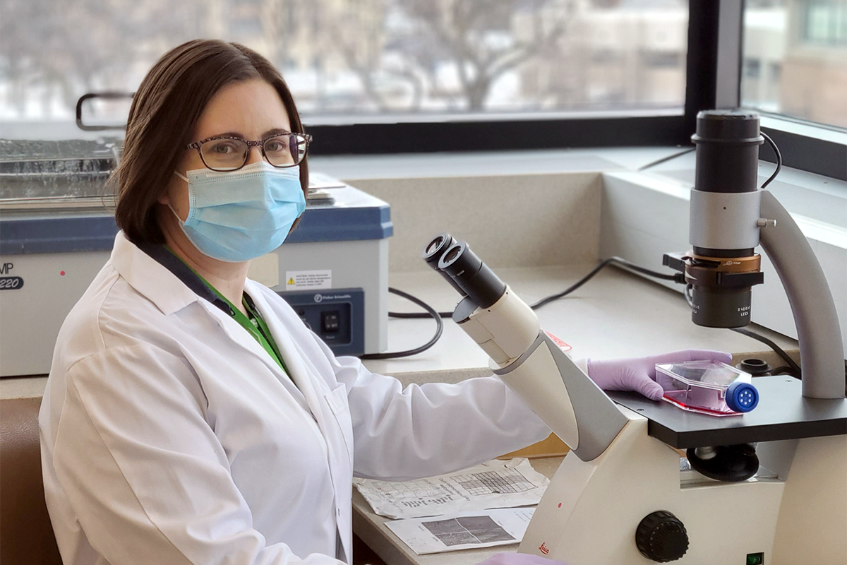 Dr. Deanna Santer, wearing a white lab coat and a surgical mask, sits in front of a microscope.