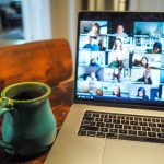 A turquoise and green coffee mug sits beside a laptop with a video chat platform on the screen