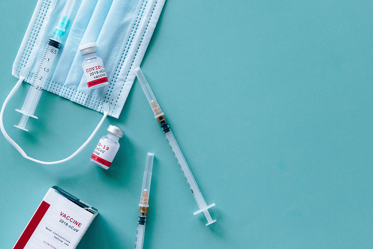 COVID-19 vaccine vials, syringes and a blue mask on a green table.