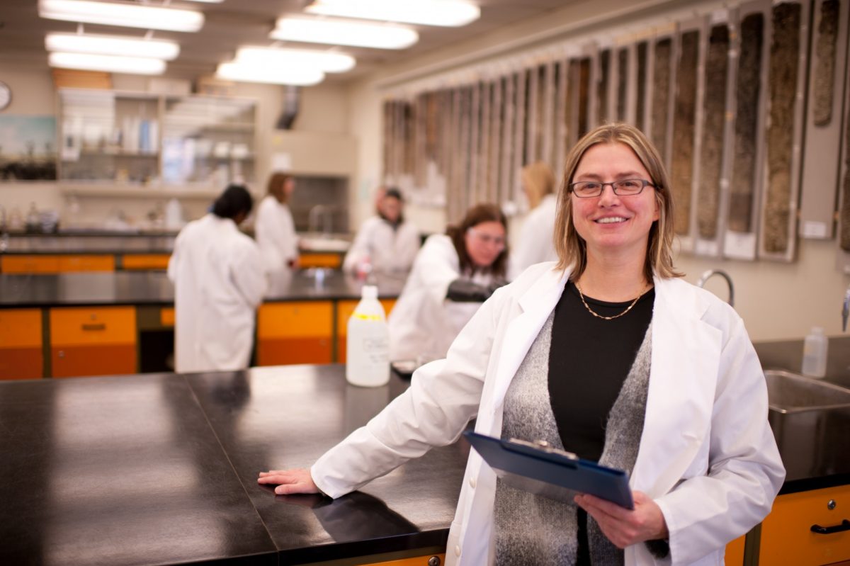 Dr. Annemieke Farenhorst wearing a lab coat, holding a clipboard, with students working in the lab behind her