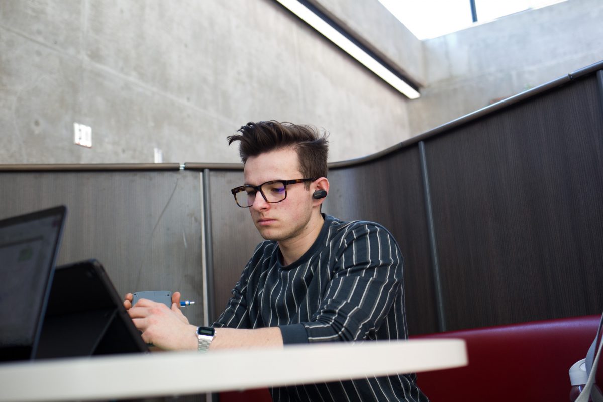 A white guy wearing glasses and a black shirt with white stripes works at a computer on campus.