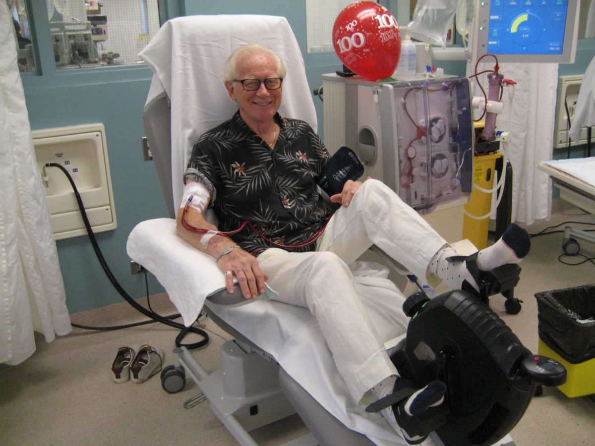 A man sitting upright in a hospital chair with his legs stretched in front of him pedals a stationary cycling wheel while receiving dialysis.