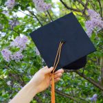 A hand holds a graduation cap in front of a lilac bush in bloom with purple flowers