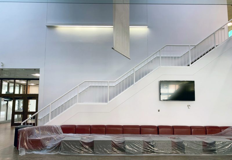 Looking at the a white staircase against a white wall in the Asper School Drake Building. Red couches are below the staircase.