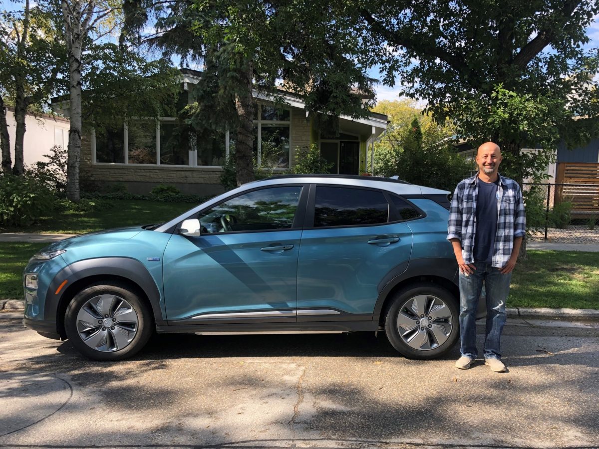 Dr. Nazim Cicek stands outside alongside his full electric vehicle, under a large tree on a sunny day.