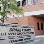 A cement sign says Drake Centre, I.H. Asper School of Business and University of Manitoba Transport Centre. It's summer and there is a green tree and the red brick Drake Building in the background.