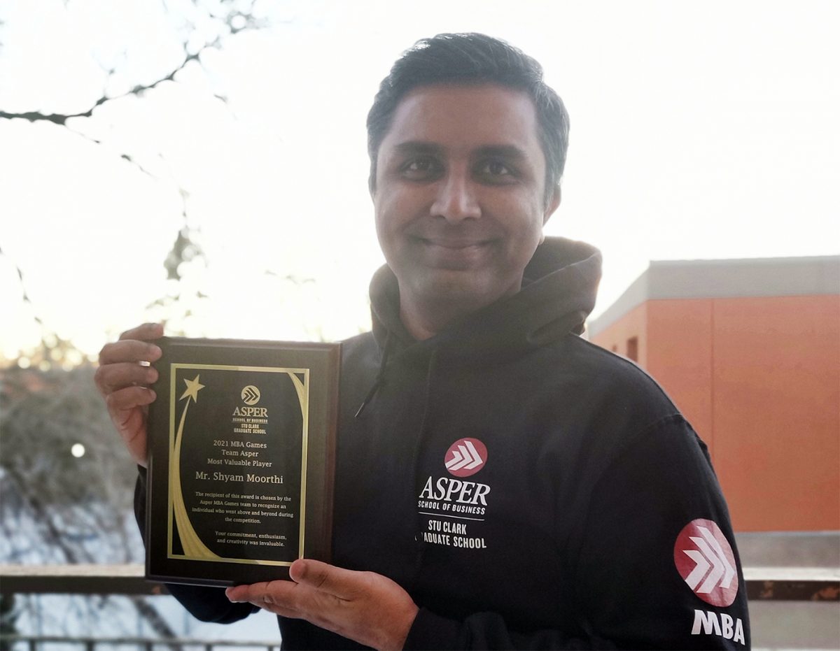 Man with dark hair holding a plaque in a black Asper School of Business hoodie.