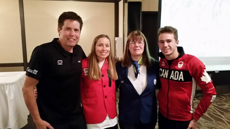 Four Olympics staff and athlete wearing red, black, and white standing together smiling. All their clothing the Canadian Olympic gear.