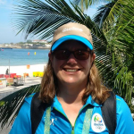 Woman wearing blue Olympics staff shirt and hat smiling with Rio beach beside her.