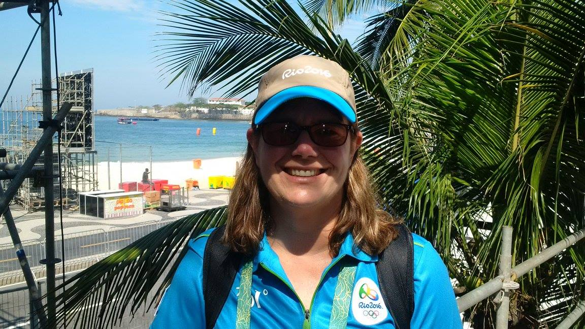 Woman wearing blue Olympics staff shirt and hat smiling with Rio beach beside her.