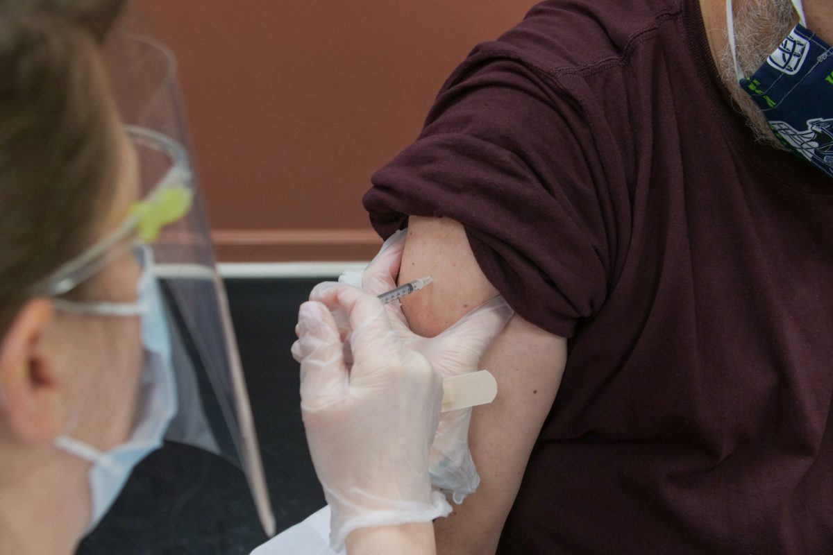 A health-care worker wearing a face shield injects a needle into a client's arm.