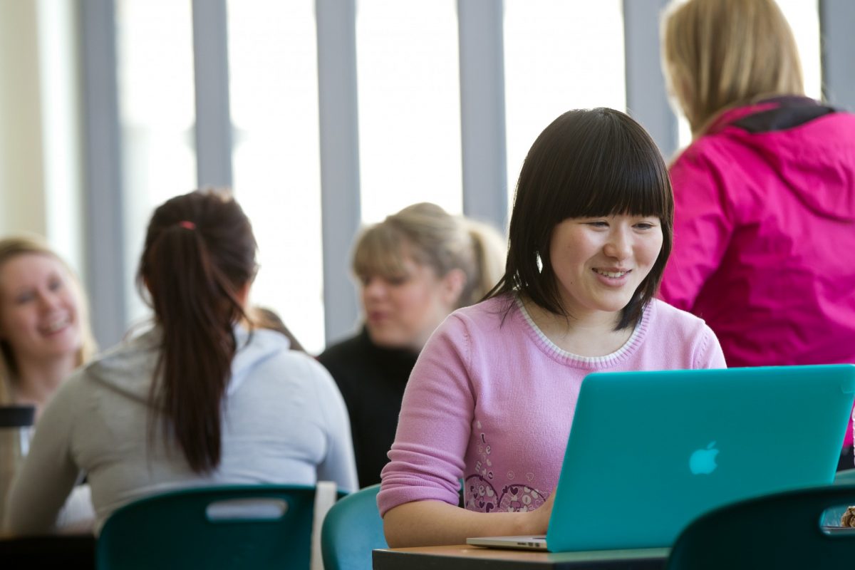 A student with dark brown hair and a pink shirt works at a turquoise laptop, surrounded by other students.