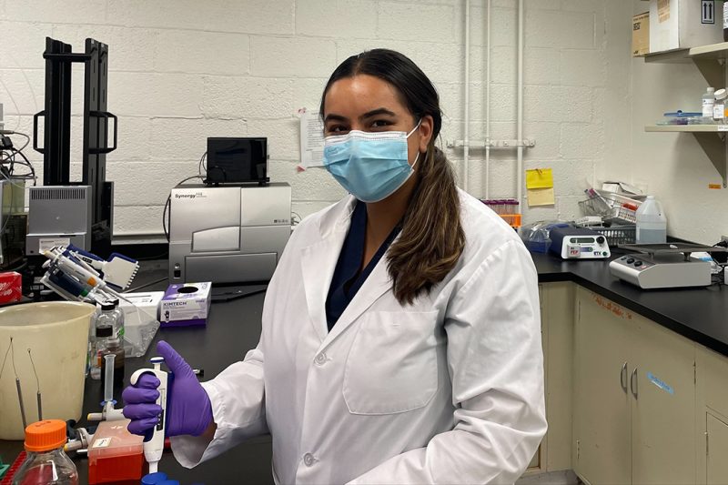 Navi Mann in a lab. She is wearing a lab coat and face mask.