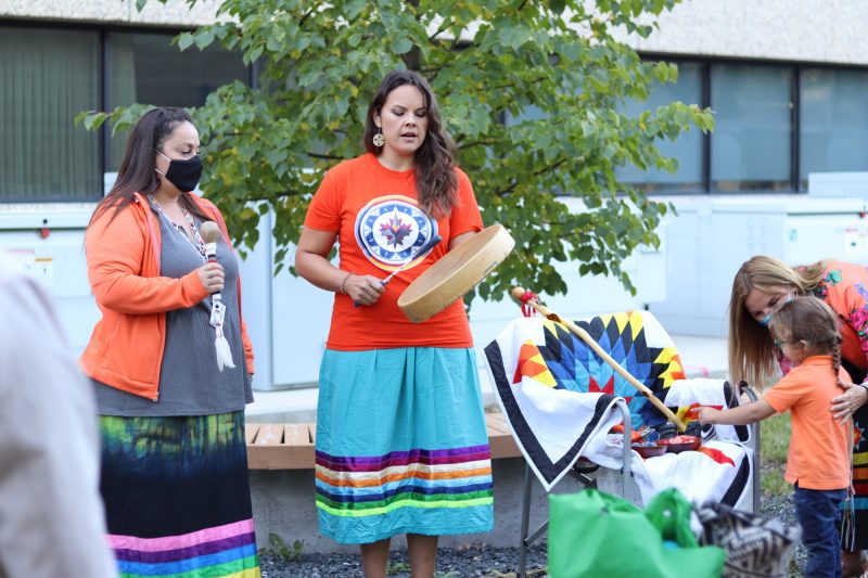 A woman wearing an orange shirt and a ribbon skirt plays a hand drum and sings at the Honouring our Children ceremony.