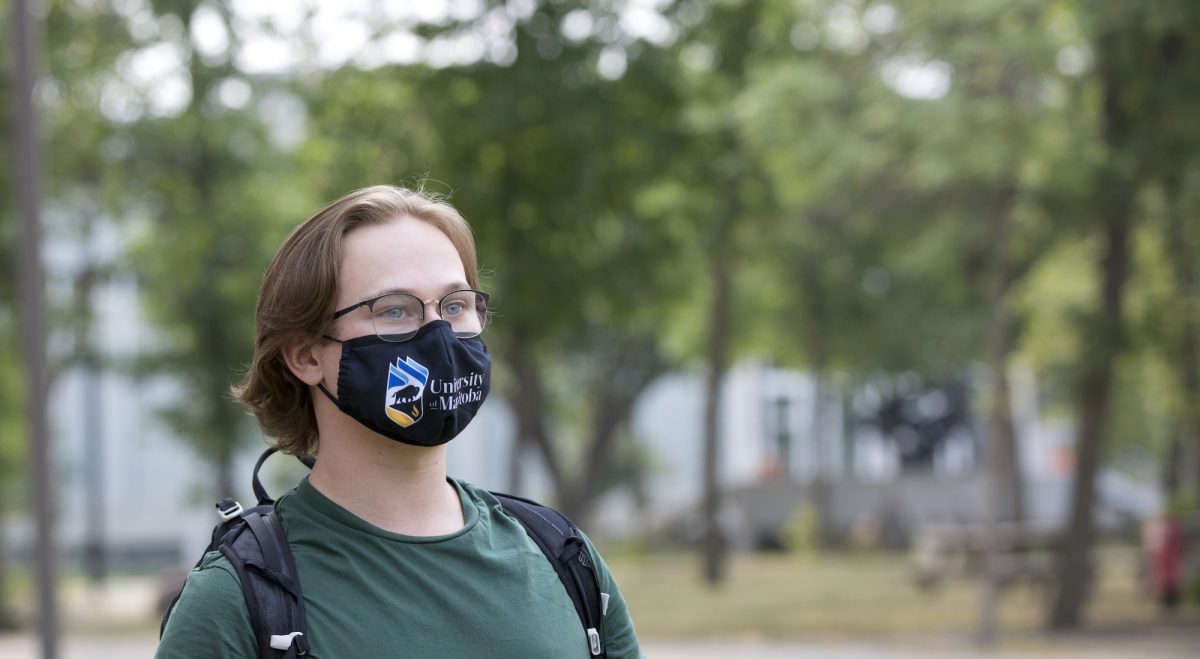 A student wearing a green shirt and a black mask with the University of Manitoba logo on it stands among the trees on the Fort Garry Campus.
