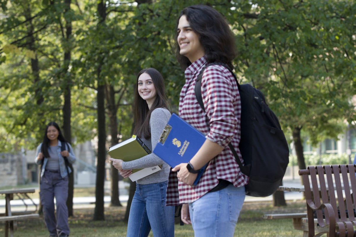 Two students smile as they walk on the Fort Garry Campus. The person in the foreground wears a red and white checked shirt with a black backpack and carries a blue UM clipboard. The person next to them wears a grey shirt and holds a couple textbooks. a