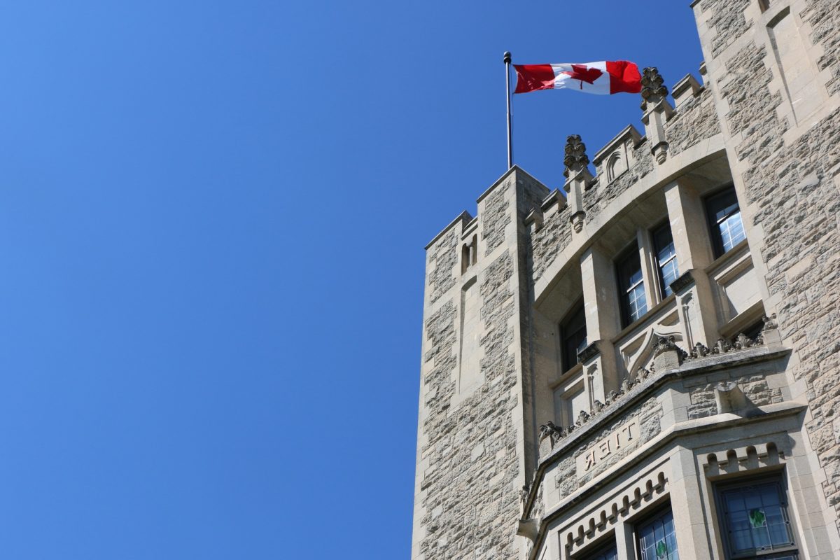 Outside of limestone building with Canadian flag on top in front of a bright blue sky.