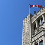 Outside of limestone building with Canadian flag on top in front of a bright blue sky.