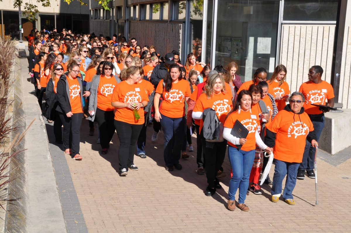 A large group of people wearing orange shirts that say Every Child Matters walk on the Fort Garry campus of the University of Manitoba.