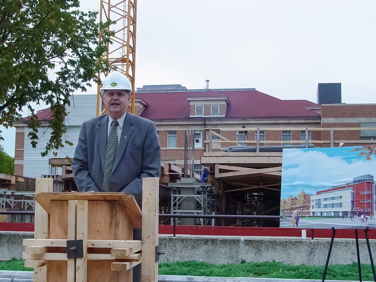Doug Ruth stands and a podium wearing a white hard hat and a suit and tie. To his left, there is a rendering of the new Engineering Information and Technology Complex.