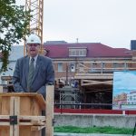 Doug Ruth stands and a podium wearing a white hard hat and a suit and tie. To his left, there is a rendering of the new Engineering Information and Technology Complex.