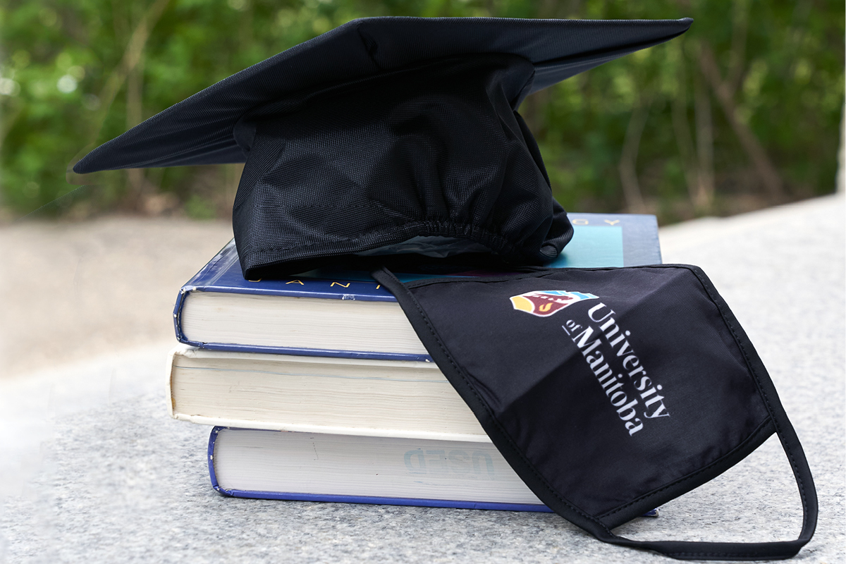 A black graduation cap and black mask with a UM logo on it sit on top of a pile of books